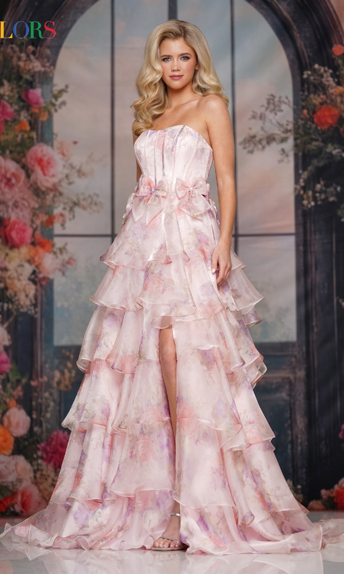 A woman poses before a floral backdrop in the Colors Dress 3809, a strapless long prom dress with a corset bodice, tiered light pink chiffon A-line skirt, floral patterns, and a high-low hem.